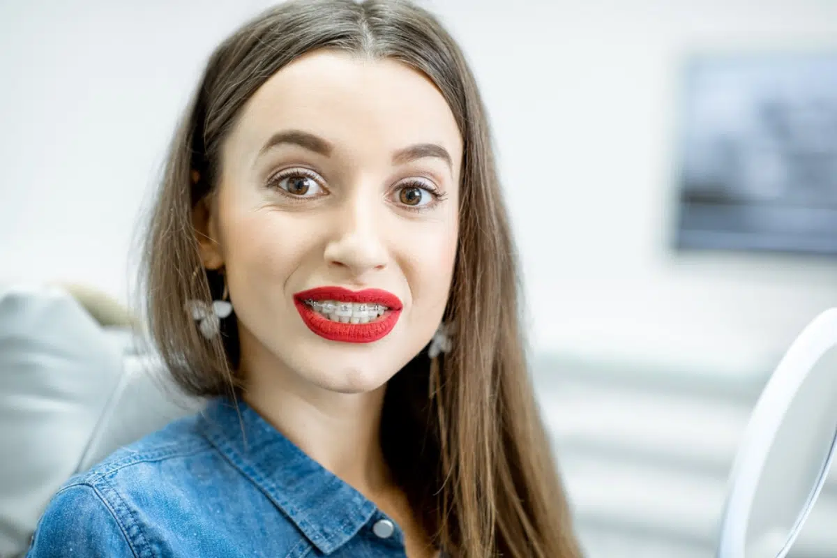 A headshot of a young woman with long brown hair, wearing red lipstick and braces, looking directly at the camera with a slight smile.
