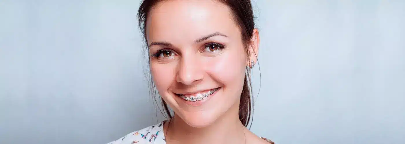 A portrait of a happy young adult woman smiling brightly at the camera, wearing subtle metal braces on her upper teeth.