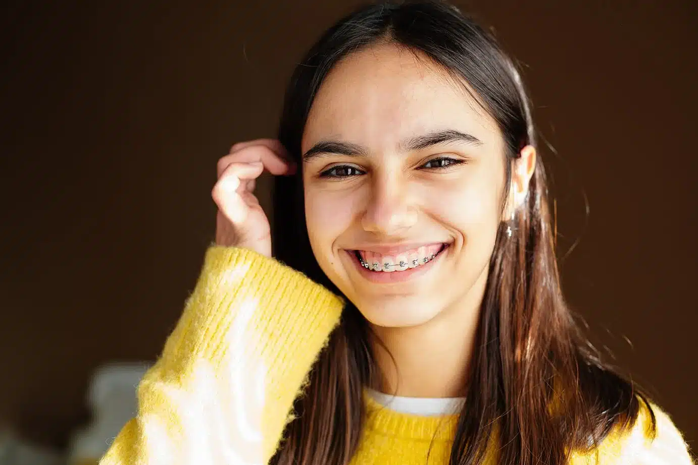 A portrait of a smiling teenage girl with long dark hair, wearing a bright yellow sweater and showing off her traditional metal braces.